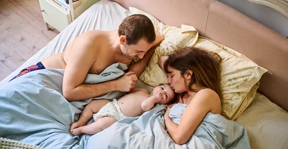 Glimlachende man en vrouw liggen samen in bed met hun jonge kind tussen hen in. De drie kijken elkaar liefdevol aan in een warme, huiselijke sfeer. Campagnebeeld voor verzekeringen 2025.